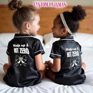 Pode incluir: Duas crian&ccedil;as vestindo pijamas de cetim preto com detalhes brancos e o texto "Wake up & HIT ZERO" nas costas. Cada parte superior do pijama tem um la&ccedil;o branco com uma estrela de cada lado.