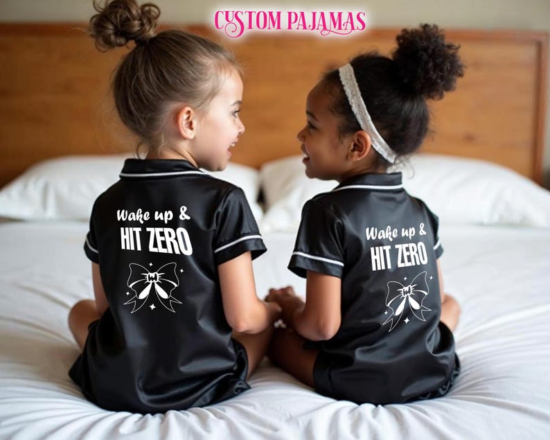 Pode incluir: Duas crian&ccedil;as vestindo pijamas de cetim preto com detalhes brancos e o texto "Wake up & HIT ZERO" nas costas. Cada parte superior do pijama tem um la&ccedil;o branco com uma estrela de cada lado.
