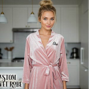 May include: A custom pink velvet robe with the name "Cleo" embroidered on the chest. The robe features a wrap-around design and a matching belt. The text "CUSTOM VELVET ROBE" is displayed at the bottom left of the image.