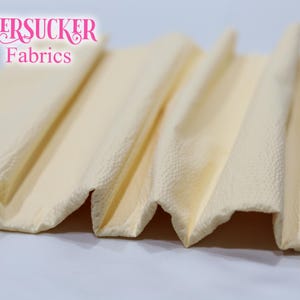 Puede incluir: Primer plano de tela plegada y texturizada en un tono amarillo p&aacute;lido. La tela tiene un aspecto de seersucker, con una superficie ligeramente arrugada. El texto "SEERSUCKER Fabrics" es visible en rosa en la parte superior.