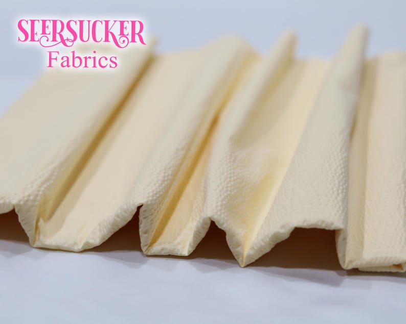 Puede incluir: Primer plano de tela plegada y texturizada en un tono amarillo p&aacute;lido. La tela tiene un aspecto de seersucker, con una superficie ligeramente arrugada. El texto "SEERSUCKER Fabrics" es visible en rosa en la parte superior.