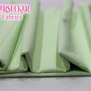 Puede incluir: Primer plano de tela seersucker verde claro doblada. La tela tiene una superficie texturizada y arrugada. Las palabras "SEERSUCKER Fabrics" est&aacute;n en escritura rosa en la esquina superior izquierda. La tela est&aacute; doblada en acorde&oacute;n.