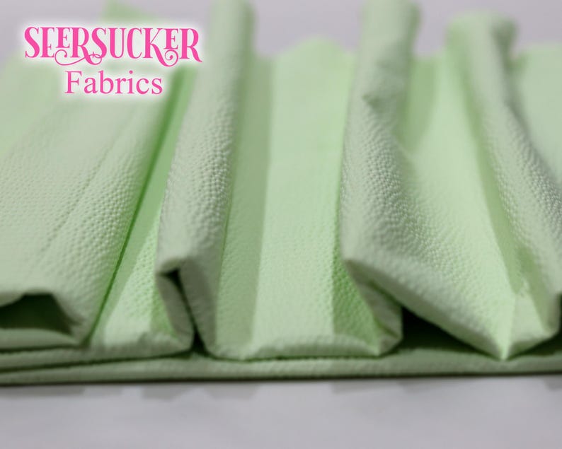 Puede incluir: Primer plano de tela seersucker verde claro doblada. La tela tiene una superficie texturizada y arrugada. Las palabras "SEERSUCKER Fabrics" est&aacute;n en escritura rosa en la esquina superior izquierda. La tela est&aacute; doblada en acorde&oacute;n.