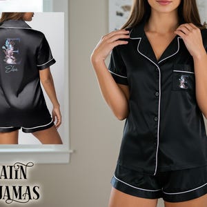 Peut inclure: Ensemble de pyjama en satin noir avec bordure blanche, comprenant une chemise boutonnée et un short. La chemise a une poche avec un motif floral initial et le nom "Elsa". Le dos de la chemise a également un motif floral initial et le nom "Elsa". Les mots "SATIN PAJAMAS" sont en bas.