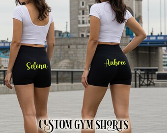 May include: Two people wearing black gym shorts with personalised names in yellow script. They are also wearing white crop tops. The words "CUSTOM GYM SHORTS" are printed in a decorative font at the bottom of the image.