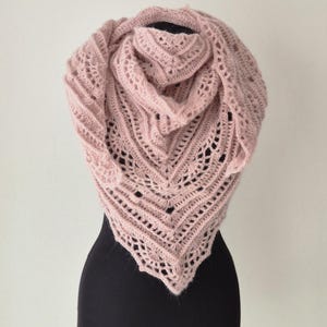 Könnte beinhalten: Ein zartes, rosa gehäkeltes Tuch mit einem Spitzenmuster. Das Tuch ist über eine schwarze Schaufensterpuppe gelegt.