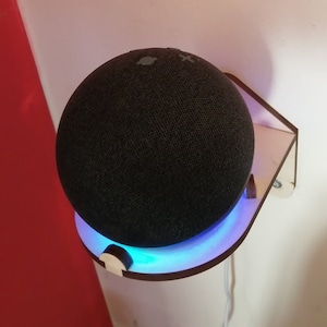 Pode incluir: Um alto-falante inteligente preto com um anel de luz azul está montado em uma prateleira de parede de madeira. A prateleira está presa a uma parede branca.