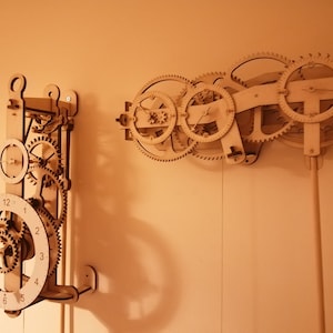 Op de afbeelding: Twee houten klokmechanismen met ingewikkelde tandwielen en wijzerplaten. De klok links heeft een ronde wijzerplaat met Romeinse cijfers en een minutenwijzer. De klok rechts heeft een complexer ontwerp met meerdere tandwielen en wijzerplaten.