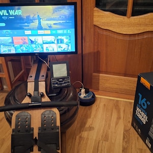 Puede incluir: Una máquina de remo de madera con un mango negro y reposapiés. La máquina tiene una pantalla digital y está conectada a una pantalla de televisión que muestra la palabra "CIVIL WAR". Una caja etiquetada "16" pulgadas wheel trims" está en el suelo.