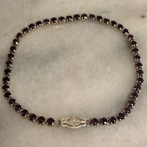 Pode incluir: Uma pulseira de ténis com uma fila contínua de pedras preciosas granada redondas, de cor vermelho-escuro, engastadas em garras de metal prateado. A pulseira inclui um fecho decorativo alongado de estilo filigrana com um motivo floral central.