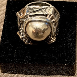 May include: A silver-toned ring featuring a large, smooth, polished spherical center element surrounded by a textured, irregular, and angular metal frame with deep grooves and sharp edges, creating a high-contrast, reflective metallic appearance.