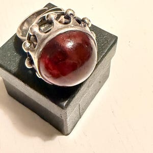 May include: A silver ring featuring a large, smooth, round, deep red cabochon stone set within a crown-shaped metal frame. The ring band is visible behind the setting, which is positioned atop a small, square, black box.