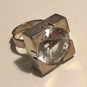 Op de afbeelding: Een zilverkleurige ring met een grote, ronde, heldere gefacetteerde steen in een vierkant metalen montuur. De ringband is verstelbaar en heeft een kleine opening aan de achterkant, waarbij het metaal een gladde, gepolijste afwerking heeft.