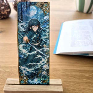 Könnte beinhalten: Ein rechteckiges Lesezeichen mit einer dunkelhaarigen Figur in dunkler Uniform, die ein Katana hält. Die Illustration zeigt wirbelnden blauen Nebel, weiße Blumen, einen Vollmond und goldfarbene geometrische Muster auf einer schmalen Karte.