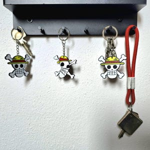 Puede incluir: Tres llaveros con un dise&ntilde;o de calavera y huesos cruzados, que llevan un sombrero de paja. Los llaveros est&aacute;n colgados de un llavero de metal negro. Un cord&oacute;n rojo con un llavero plateado tambi&eacute;n est&aacute; colgado del llavero.