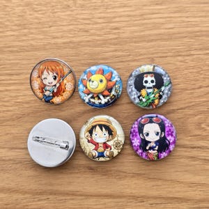 Könnte beinhalten: Sechs runde Ansteckbuttons mit farbenfrohen Anime-Charakter-Illustrationen. Fünf Buttons zeigen Chibi-Figuren, darunter ein Mädchen mit orangefarbenen Haaren, ein sonnenartiges Wesen, ein Skelett-Musiker, ein Junge mit Strohhut und ein schwarzhaariges Mädchen.