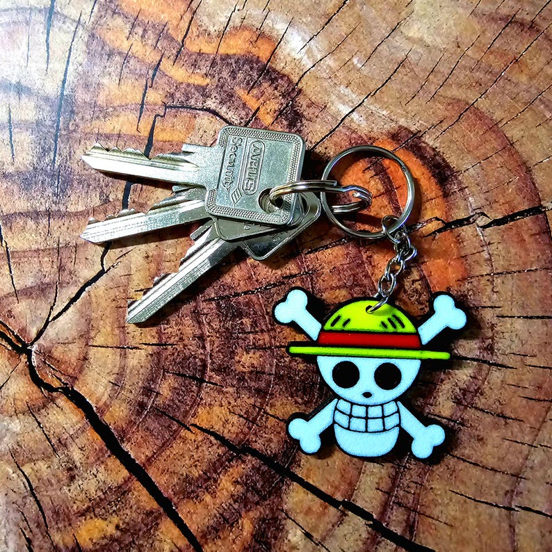 Puede incluir: Un llavero plateado con un dije de calavera y huesos cruzados blancos. La calavera lleva un sombrero de paja amarillo y rojo. El llavero est&aacute; unido a un juego de llaves con una etiqueta de seguridad AVENTIS plateada.
