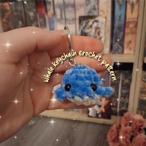 Könnte beinhalten: Ein blau-weiß gehäkelter Wal-Schlüsselanhänger mit schwarzen Augen und einem silbernen Schlüsselring. Der Text "whale keychain crochet pattern" ist in einer geschwungenen Schrift um den Wal geschrieben.