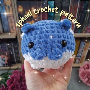 Könnte beinhalten: Handgefertigtes blau-weißes Häkeltier mit schwarzen Augen und kleinen Ohren. Der Text "spheal crochet pattern" ist in einer Schreibschrift geschrieben. Das Kuscheltier wird in einer Hand gehalten.