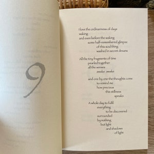 Pu&ograve; includere: Libro aperto con una poesia stampata sulla pagina di destra. La poesia &egrave; in carattere nero e la pagina di sinistra ha un simbolo grigio del numero nove. Il libro &egrave; su una superficie di legno chiaro.