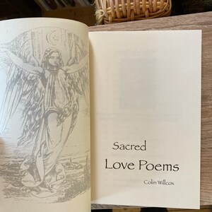 Pu&ograve; includere: Libro aperto con il titolo "Sacred Love Poems" di Colin Willcox. La pagina sinistra presenta un'illustrazione dettagliata di un angelo con le ali spiegate, una luna crescente e una stella. Le pagine del libro sono bianco sporco.