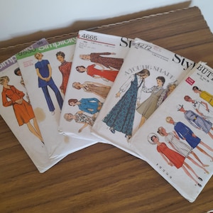 May include: A selection of vintage sewing pattern envelopes, featuring illustrations of women's clothing. The patterns include dresses, jumpsuits, and coats. Brands include Simplicity and Butterick. The envelopes are displayed on a wooden surface.