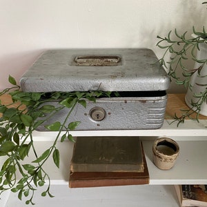 May include: A vintage metal cash box with a lock and key, sitting on a white shelf with books and a small ceramic pot. The box is surrounded by green leafy plants.