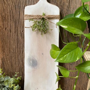 May include: A rectangular wooden cutting board with a white-washed finish and a handle at the top. The board features a natural wood grain visible through the paint, a dark knot, and a small bundle of green sprigs tied with brown twine.