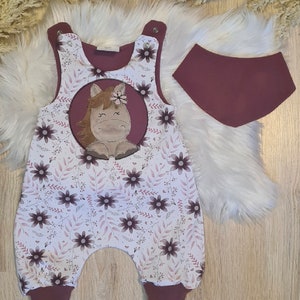 Könnte beinhalten: Ein weißer und brauner Baby-Strampler mit Blumenmuster und einem braunen Pferde-Applikationen. Der Strampler hat braune Träger und braune Einfassungen an den Beinen. Ein braunes Lätzchen ist ebenfalls enthalten.