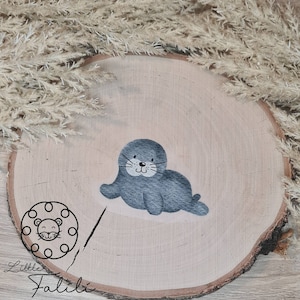 Könnte beinhalten: Ein rundes Holzschild mit einer grauen Robbenillustration und dem Text "Little Falibi" in schwarzen Buchstaben.