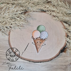 Könnte beinhalten: Ein handbemaltes rundes Schild aus Holz mit einem niedlichen Comic-Eiswaffel mit drei Kugeln Eis. Die Eiswaffel hat ein lächelndes Gesicht und hält die Arme hoch. Die Eiskugeln sind grün, orange und lila. Das Schild trägt den Text "Little Falili" auf sich.