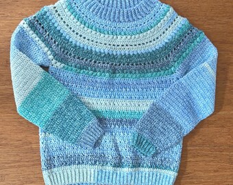 Op de afbeelding: Een handgemaakte, blauwe gehaakte trui met een ronde hals en lange mouwen. De trui heeft een kleurengradatie, die overgaat van lichtblauw naar turquoise en donkerdere blauwtinten. De mouwen hebben een gestreept patroon en de body heeft een getextureerde steek.
