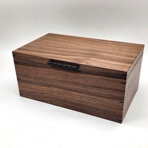 May include: A rectangular wooden box with a dark brown finish and a black handle. The box is made of wood with visible grain patterns and has a hinged lid. The box is likely used for storage or display.