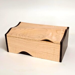 May include: A wooden box with a light-coloured, wavy top and dark brown sides. The box has a rectangular shape with a curved design on the lid. The wood grain is visible, adding texture to the piece. The box is likely used for storage or display.
