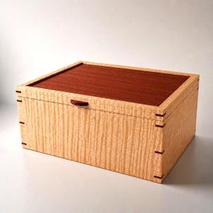 May include: A rectangular wooden box featuring a light-coloured, wavy-grained wood body with a contrasting dark reddish-brown wood lid. The box includes small dark wood accents at the corner joints and a small, semi-circular dark wood handle.
