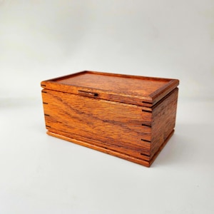 May include: A rectangular wooden box with a hinged lid. The box is made of reddish-brown wood, with visible wood grain and dovetail joints at the corners. The box is closed and sits on a white surface.