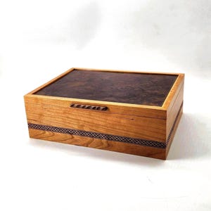 May include: A rectangular wooden box featuring a dark brown burl wood lid with a natural grain pattern. The box body is crafted from light-colored wood and includes a horizontal decorative band with a dark, intricate braided pattern.