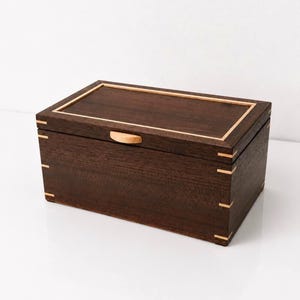 May include: A rectangular dark brown wooden box with a hinged lid. The box features light-coloured wood inlay accents along the top edges, corners, and a small semi-circular handle on the front of the lid, showcasing visible wood grain.
