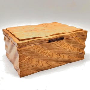 May include: A handcrafted wooden box with a wavy, textured design. The box is made of light brown wood with a darker brown handle. The lid has a unique, uneven edge. The box is likely used for storing jewelry or other small items.