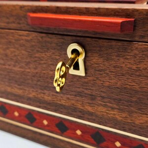 May include: A dark brown wooden box featuring a horizontal red accent strip along the top and a decorative band with red and black diamond patterns at the bottom. A gold-coloured metal key is inserted into a matching gold-coloured keyhole plate.