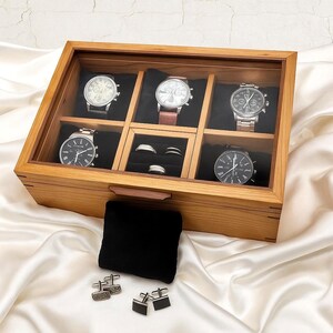 May include: A wooden watch display case with a glass lid, displaying six watches with different strap colours and face designs. The box also contains a black velvet pad, cufflinks, and rings. The box is made of light-coloured wood.