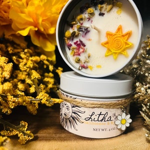 Puede incluir: Una vela blanca en una lata de metal, adornada con flores secas y un sol amarillo. La lata está etiquetada como "Litha" con un gráfico de sol y un dije de margarita. La vela pesa 113 gramos.