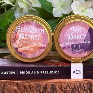 Może przedstawiać: Dwa okrągłe puszki ze złotymi obwódkami i etykietami. Na jednej napisano "Elizabeth Bennet, Elderberry Nectar", na drugiej "Mr. Darcy, Black Coral & Moss". Leżą na książce "Pride and Prejudice" Jane Austen.
