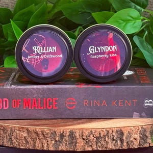 May include: Two black-lidded containers with labels reading "Killian Amber & Driftwood" and "Glyndon Raspberry Kiss" sit atop a book titled "God of Malice" by Rina Kent. The items rest on a wooden surface with green foliage in the background.