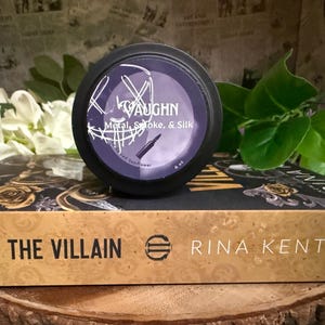 May include: A black container with a purple label that reads "VAUGHN Metal, Smoke, & Silk" sits atop a book titled "HUNT THE VILLAIN" by Rina Kent. The background includes white flowers and green leaves, with a newspaper print.
