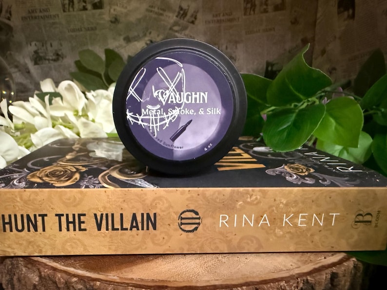 May include: A black container with a purple label that reads "VAUGHN Metal, Smoke, & Silk" sits atop a book titled "HUNT THE VILLAIN" by Rina Kent. The background includes white flowers and green leaves, with a newspaper print.