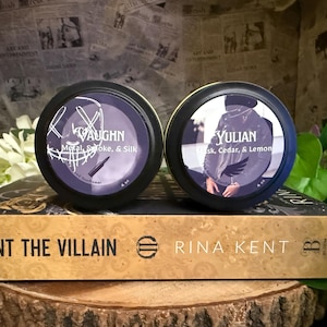 May include: Two round, black-rimmed containers with purple labels, one labeled "VAUGHN" and the other "YULIAN." The "VAUGHN" container lists "Metal, Smoke, & Silk," and the "YULIAN" container lists "Musk, Cedar, & Lemon." A book titled "HUNT THE VILLAIN" is below.