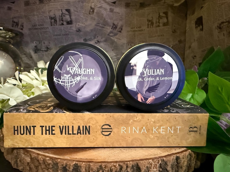 May include: Two round, black-rimmed containers with purple labels, one labeled "VAUGHN" and the other "YULIAN." The "VAUGHN" container lists "Metal, Smoke, & Silk," and the "YULIAN" container lists "Musk, Cedar, & Lemon." A book titled "HUNT THE VILLAIN" is below.