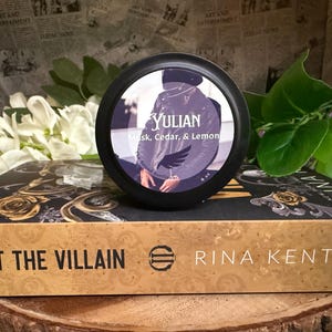 May include: A black container with the word "YULIAN" and the scent description "Musk, Cedar, & Lemon" sits atop a book titled "HUNT THE VILLAIN" by Rina Kent. The book has gold floral designs. The background includes white flowers and greenery.