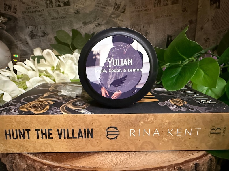 May include: A black container with the word "YULIAN" and the scent description "Musk, Cedar, & Lemon" sits atop a book titled "HUNT THE VILLAIN" by Rina Kent. The book has gold floral designs. The background includes white flowers and greenery.